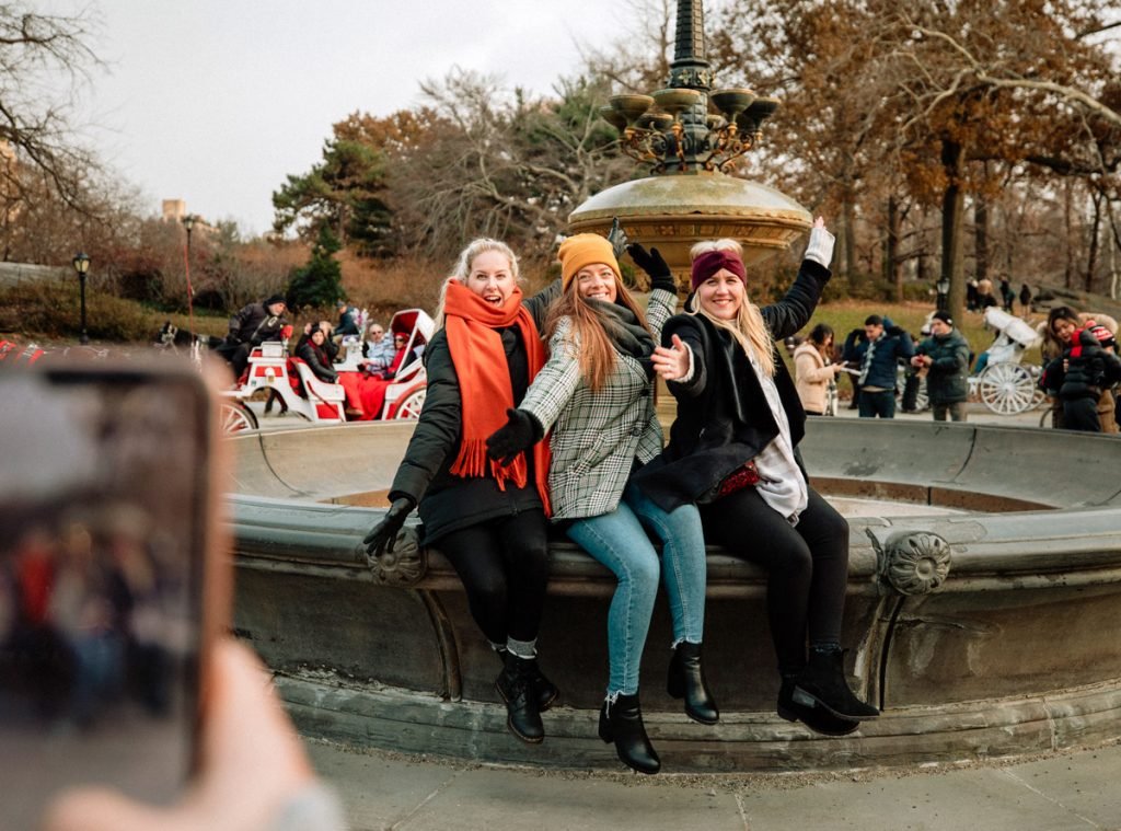 Women pose for a photo in front of a fountain in Central Park. They are wearing winter clothes. 