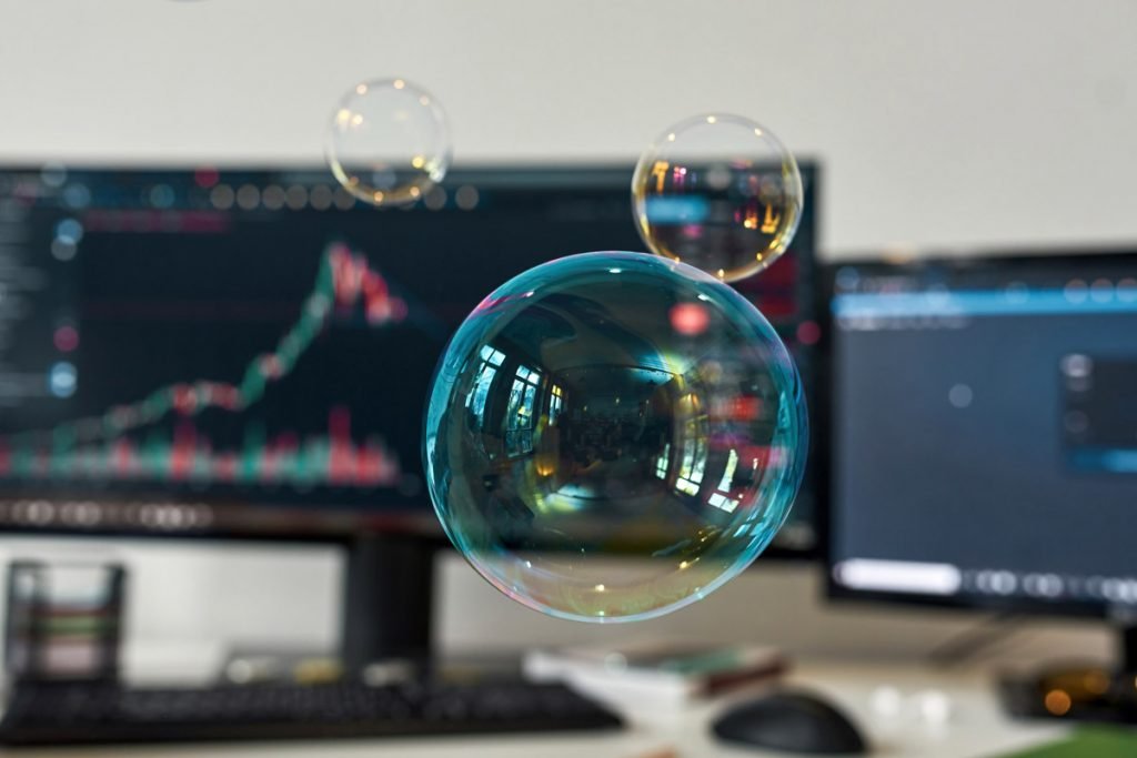 Bubbles appear in focus with a computer showing the stock market behind it. 
