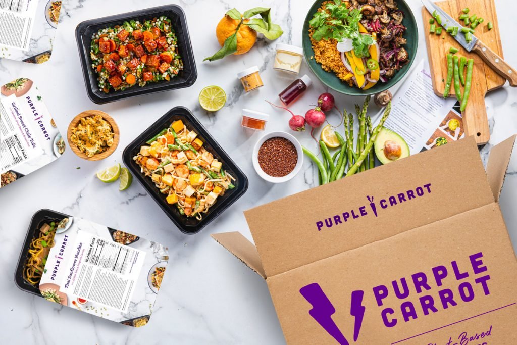 A purple carrot box is photographed with prepared vegan meals sprouting from the box.