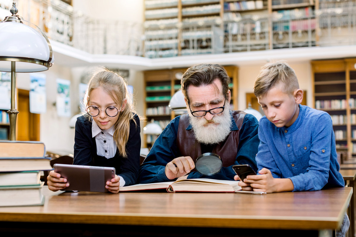 A man looks at a book in the library with his two grandchildren. 