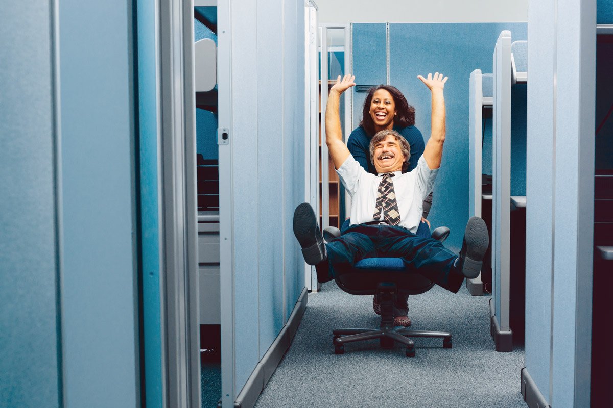 Two cubicle workers push each other on a chair down the hallway at their office. 