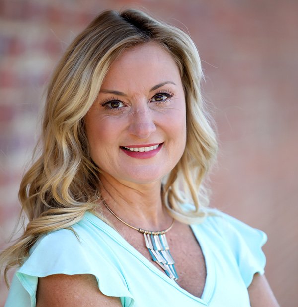 A portrait of a CEO work from home company, Tricia Sciortino. She smiling while leaning against a brick wall, and she's wearing a blue shirt.