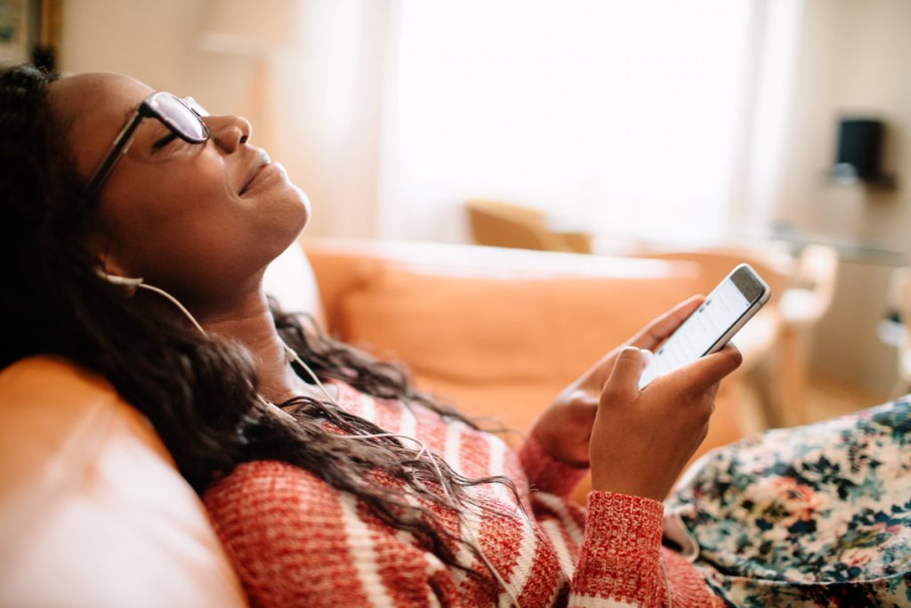 A woman reclines on her couch while listening to a podcast and smiling with her eyes closed. 