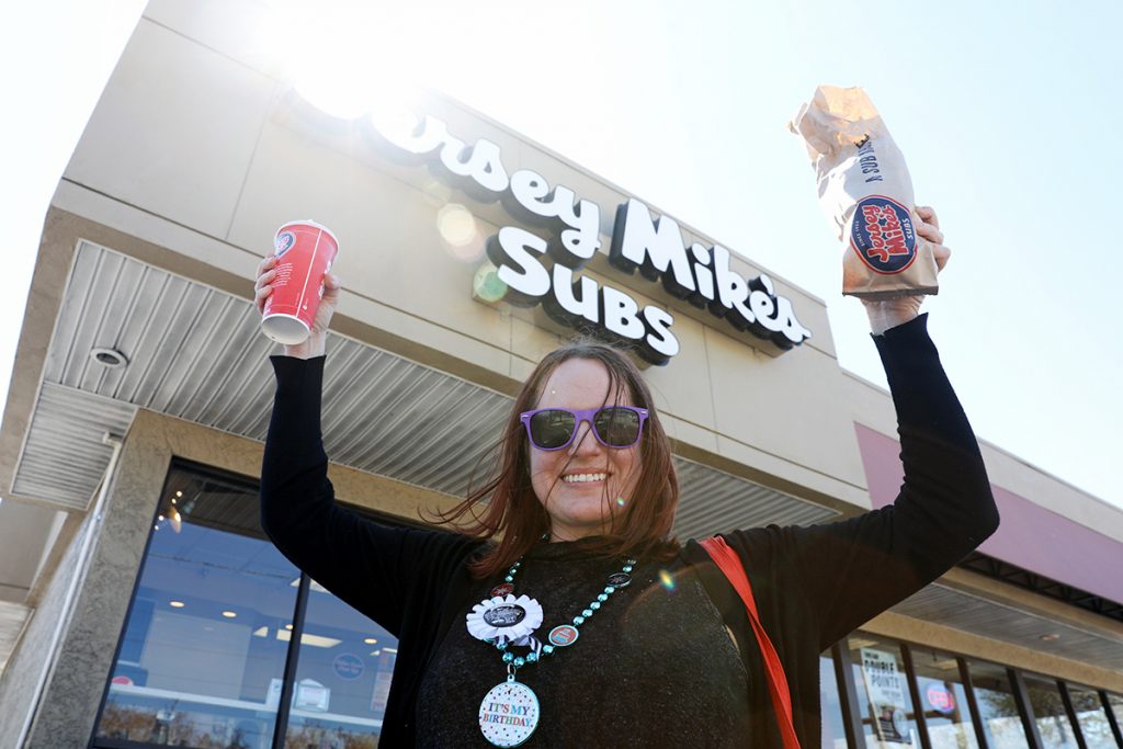 A woman smiles as she holds up a drink and a sub she got for free from Jersey Mike's Subs.