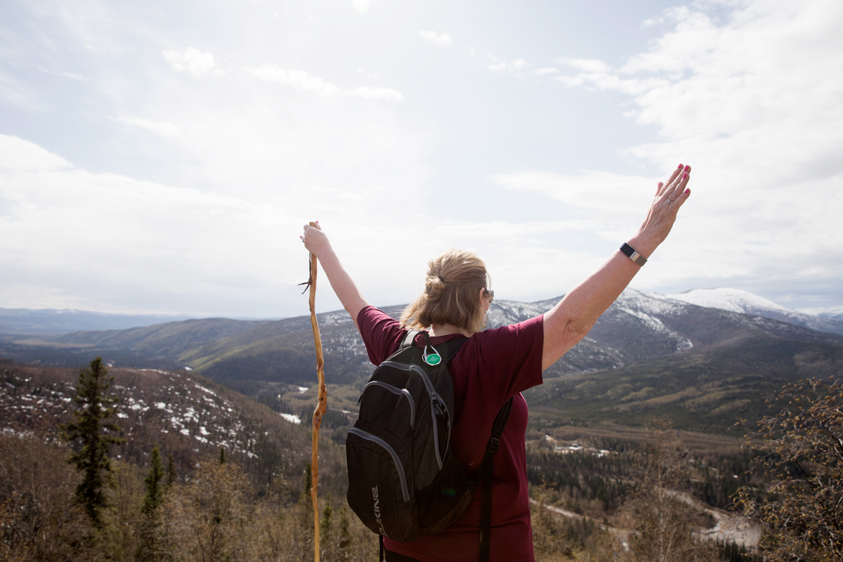 A woman waves her hands in the air as she overlooks a mountainous view in Alaska.